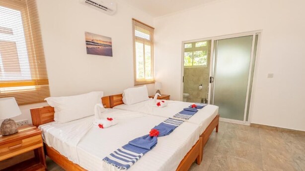 A double bed with white linens and a blue and white striped runner, a wooden nightstand with a lamp, a painting on the wall, and a sliding glass door leading to a balcony. - MacMillan's Holiday Villas await you offering a modern living space (Grand Anse Praslin)