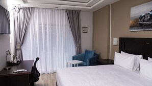 A hotel room with a large bed, a desk with a chair, a small table, a blue armchair, and a painting on the wall. - Olive Comfort Boutique Luxury Suites (Kwekwe)