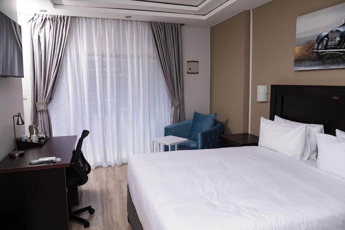 A hotel room with a large bed, a desk with a chair, a small table, a blue armchair, and a painting on the wall.