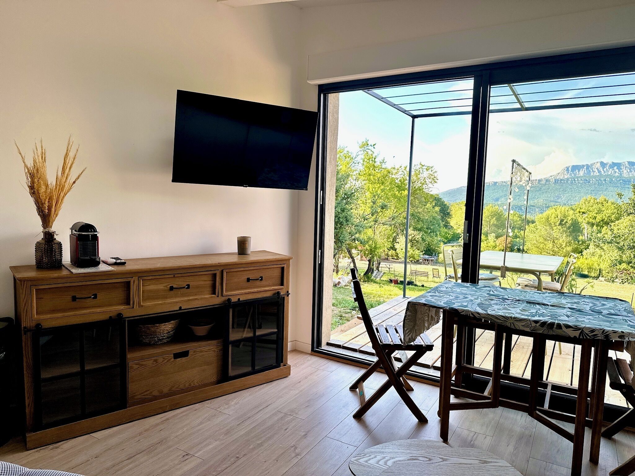 A modern living room with a large window, wooden furniture, and a flat-screen TV mounted on the wall.