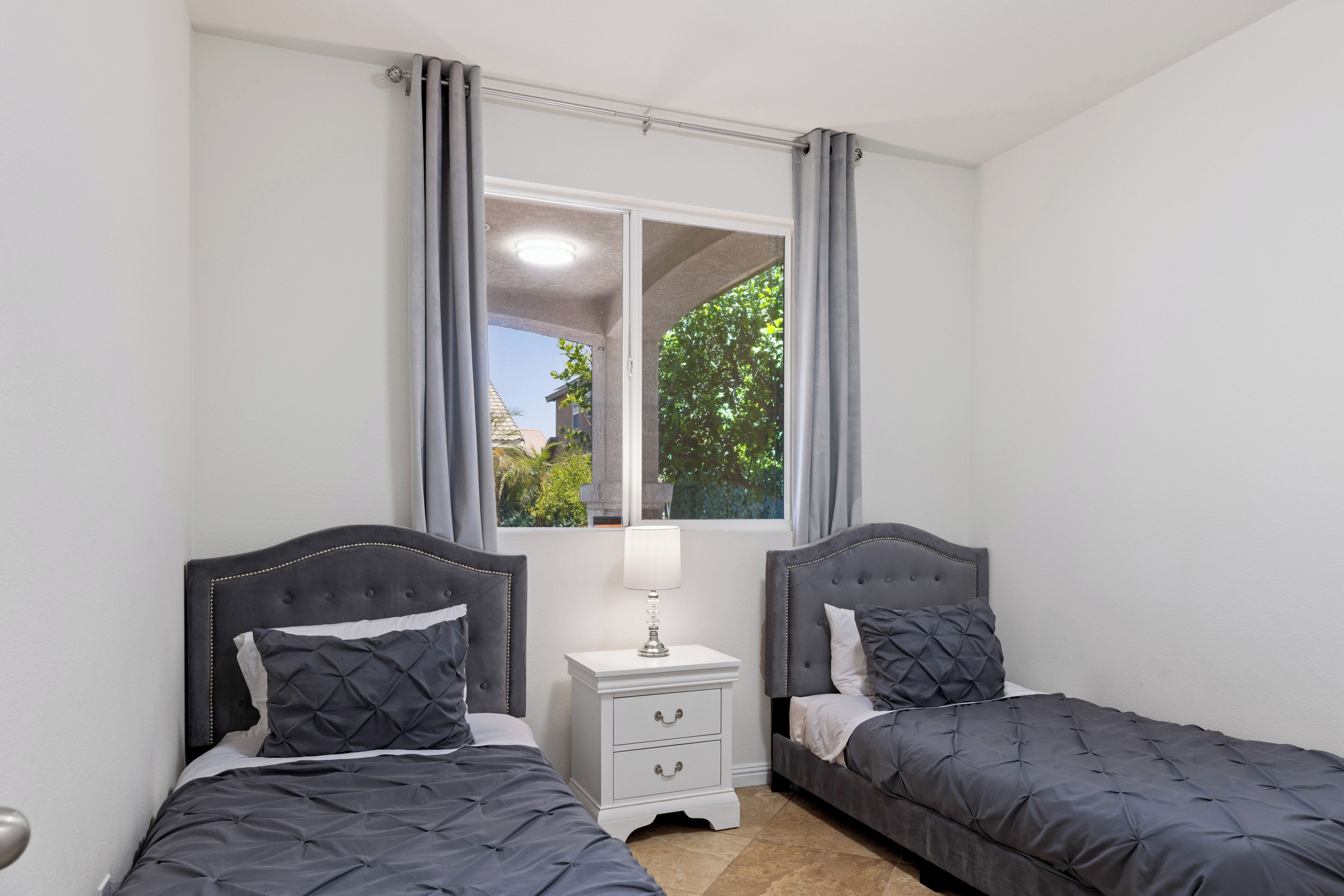 A bedroom with two beds, a nightstand with drawers, a window with curtains, and a view of greenery outside.