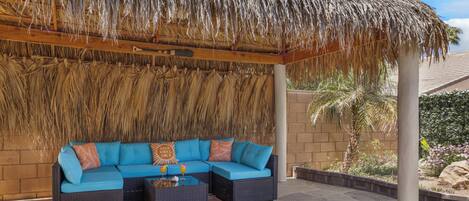 A covered outdoor seating area with a thatched roof, a sectional sofa, and a small table with drinks.