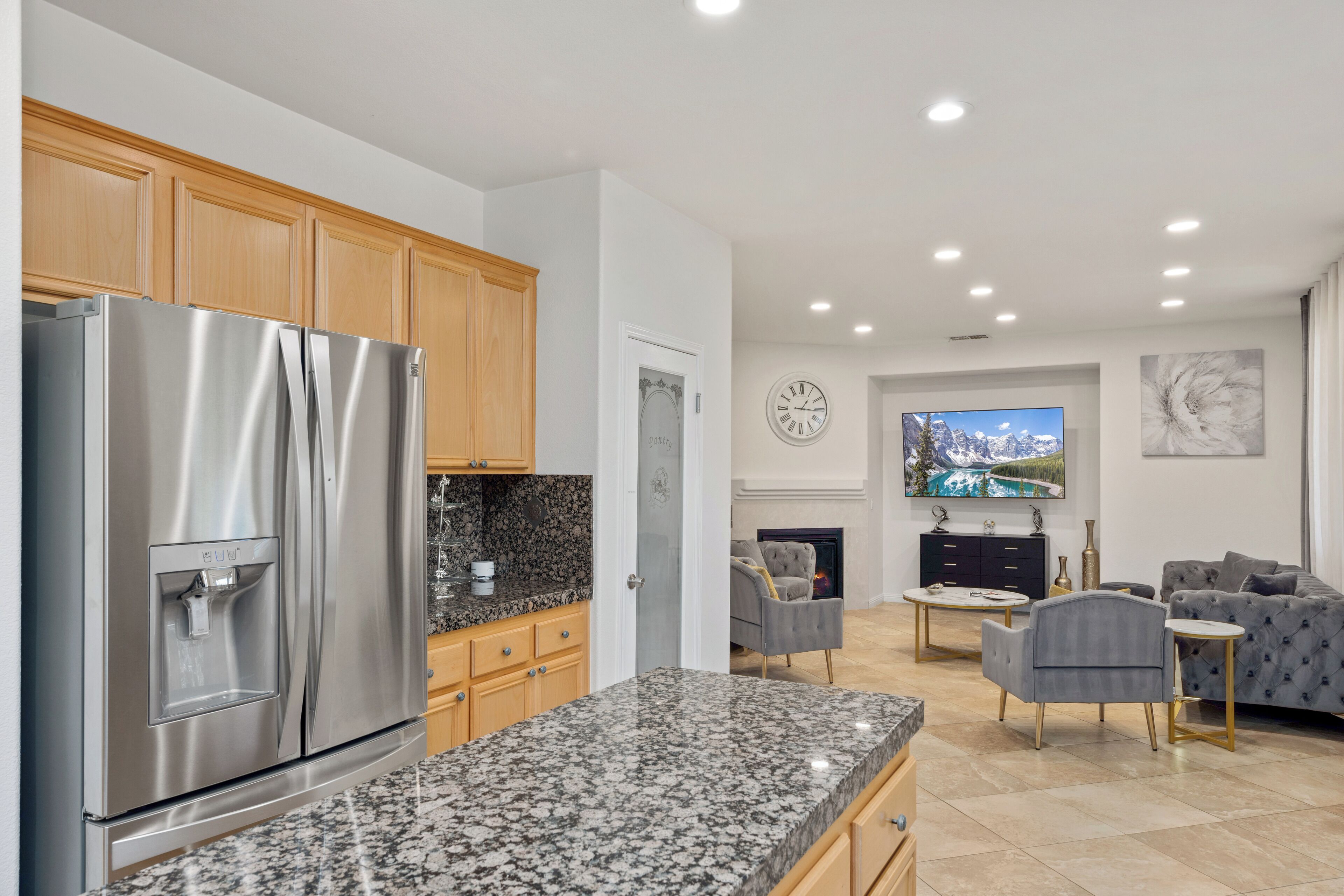 A modern kitchen with a stainless steel refrigerator, granite countertops, wooden cabinets, and a dining area with a gray sofa and coffee table.