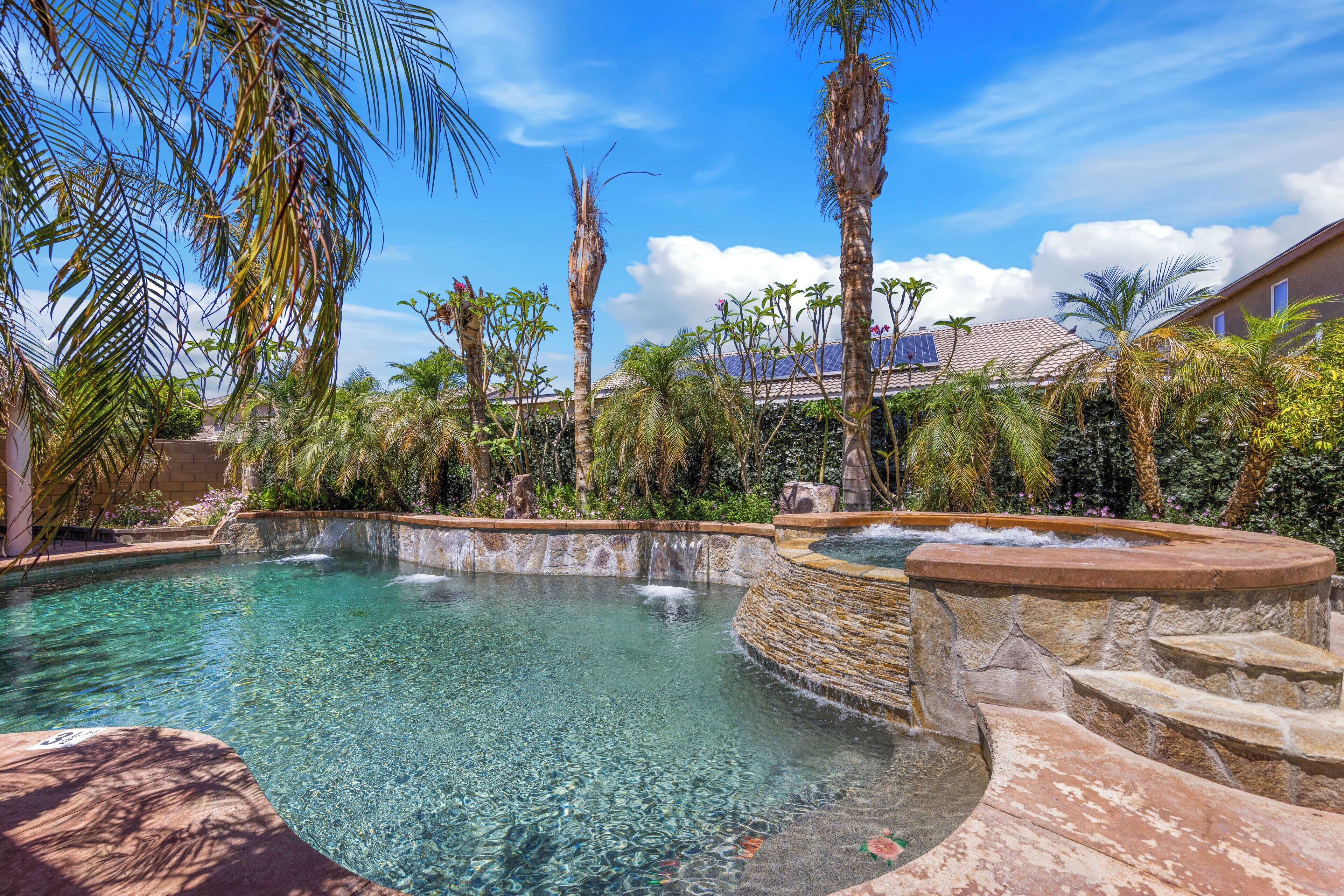 A clear pool with a waterfall feature, surrounded by palm trees and a stone wall.