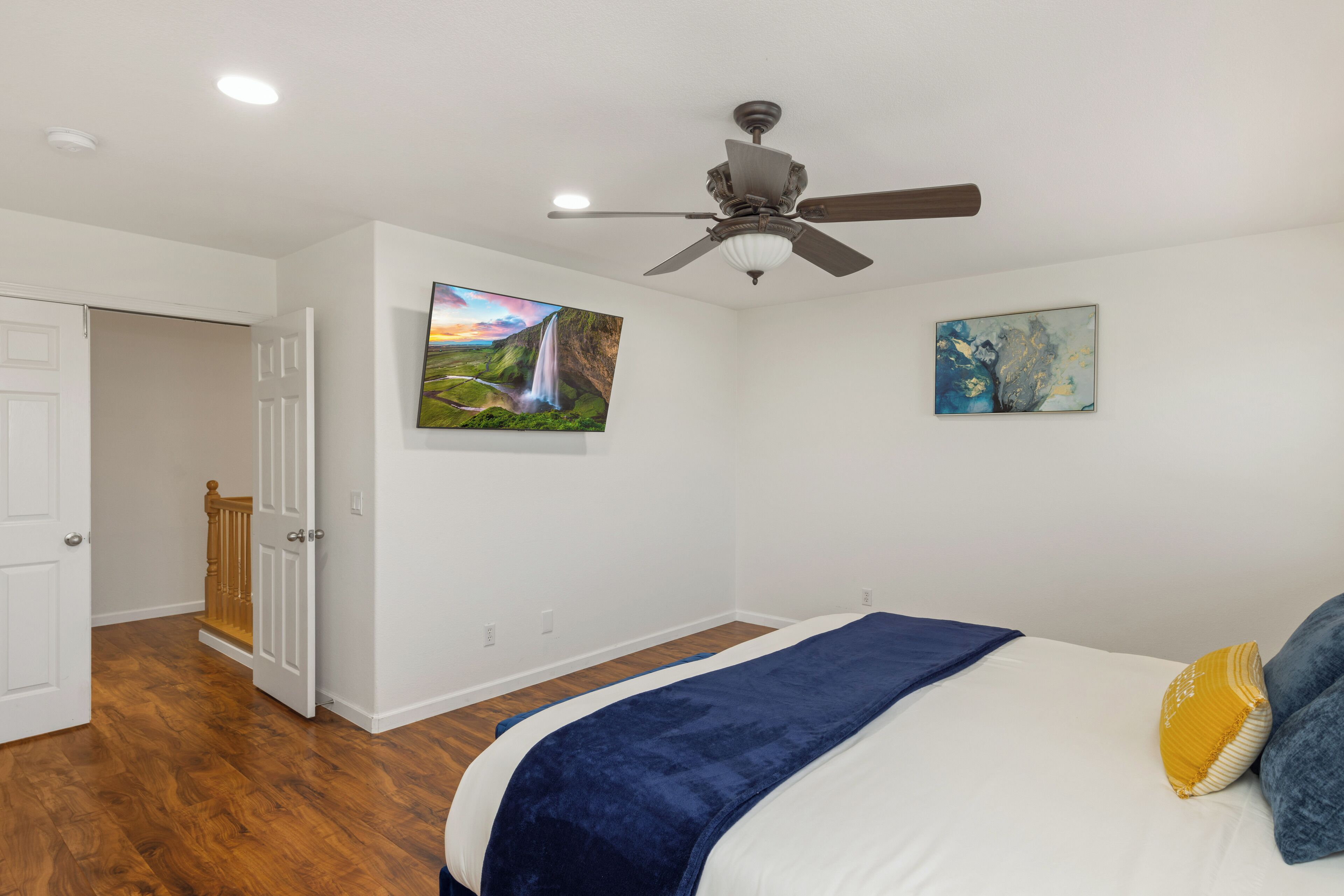 A bedroom with a bed, a ceiling fan, wooden flooring, and two paintings on the wall.
