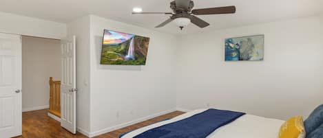 A bedroom with a bed, a ceiling fan, wooden flooring, and two paintings on the wall.