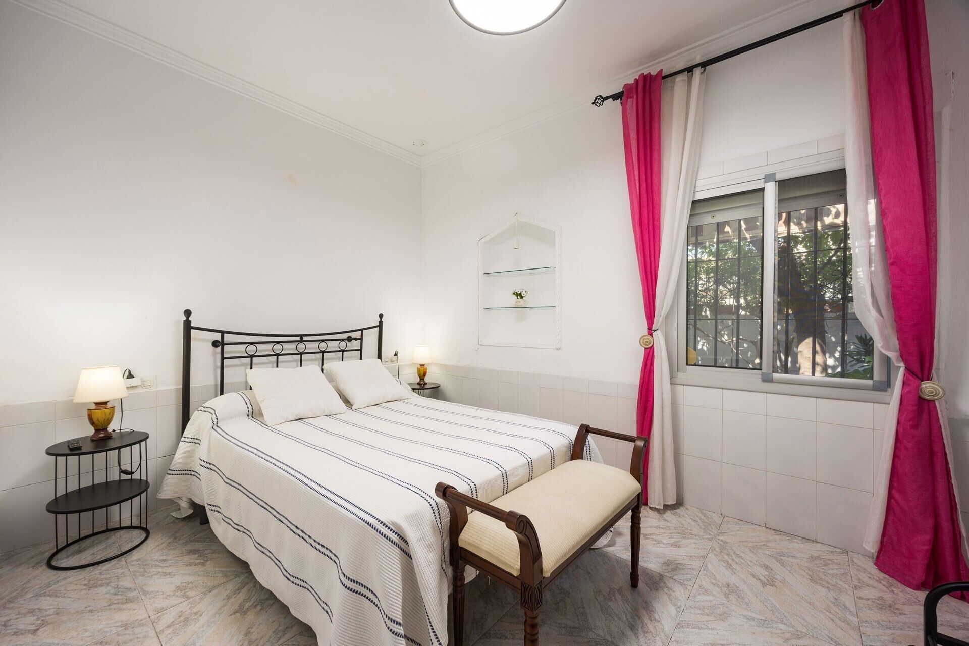 Ein Schlafzimmer mit einem Bett, Nachttischen, einem Fenster mit Vorhängen und einer kleinen Ablagefläche.