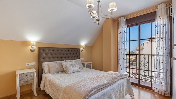 A bedroom with a tufted headboard, a large bed with a beige bedspread, two bedside tables with lamps, and a view through a glass door.