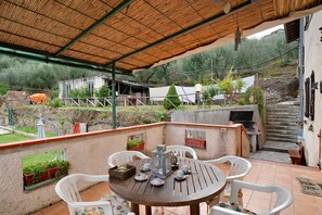 A covered patio with a table set for dining, surrounded by white chairs, and a view of a rustic house with a staircase leading to a higher level. - Apartment "Câ de Bacî" with Private Terrace (Vessalico)