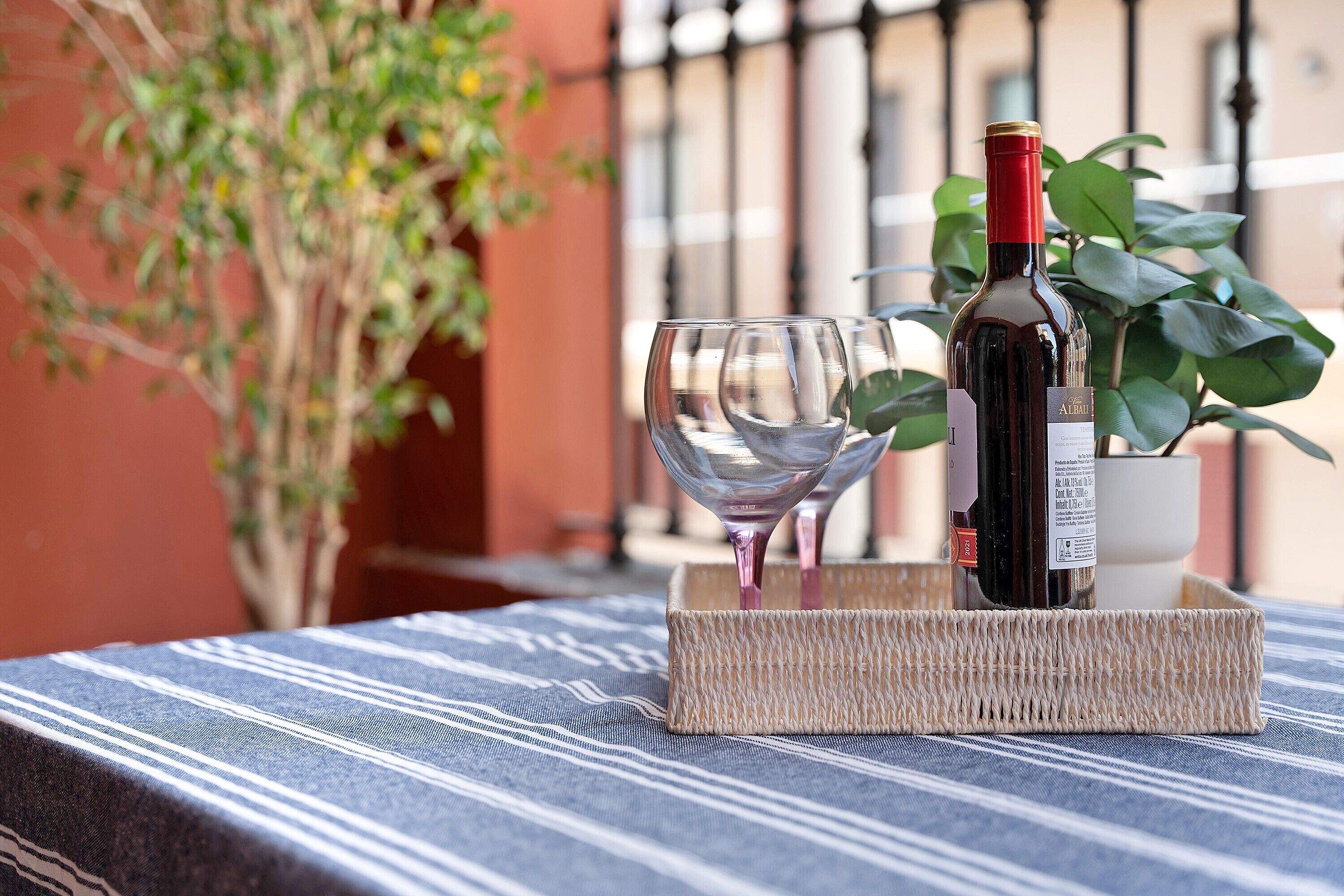 Une bouteille de vin et un verre sur une table recouverte d’une nappe à rayures, un plateau en osier et une plante en pot en arrière-plan.