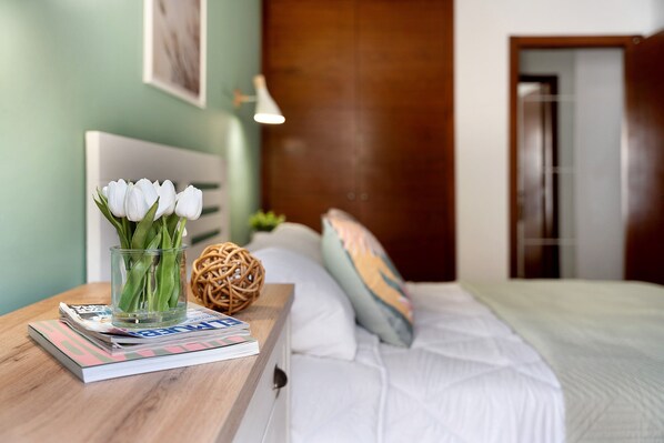 A neatly made bed with white bedding, a glass vase with white flowers, and a magazine on a wooden bedside table.