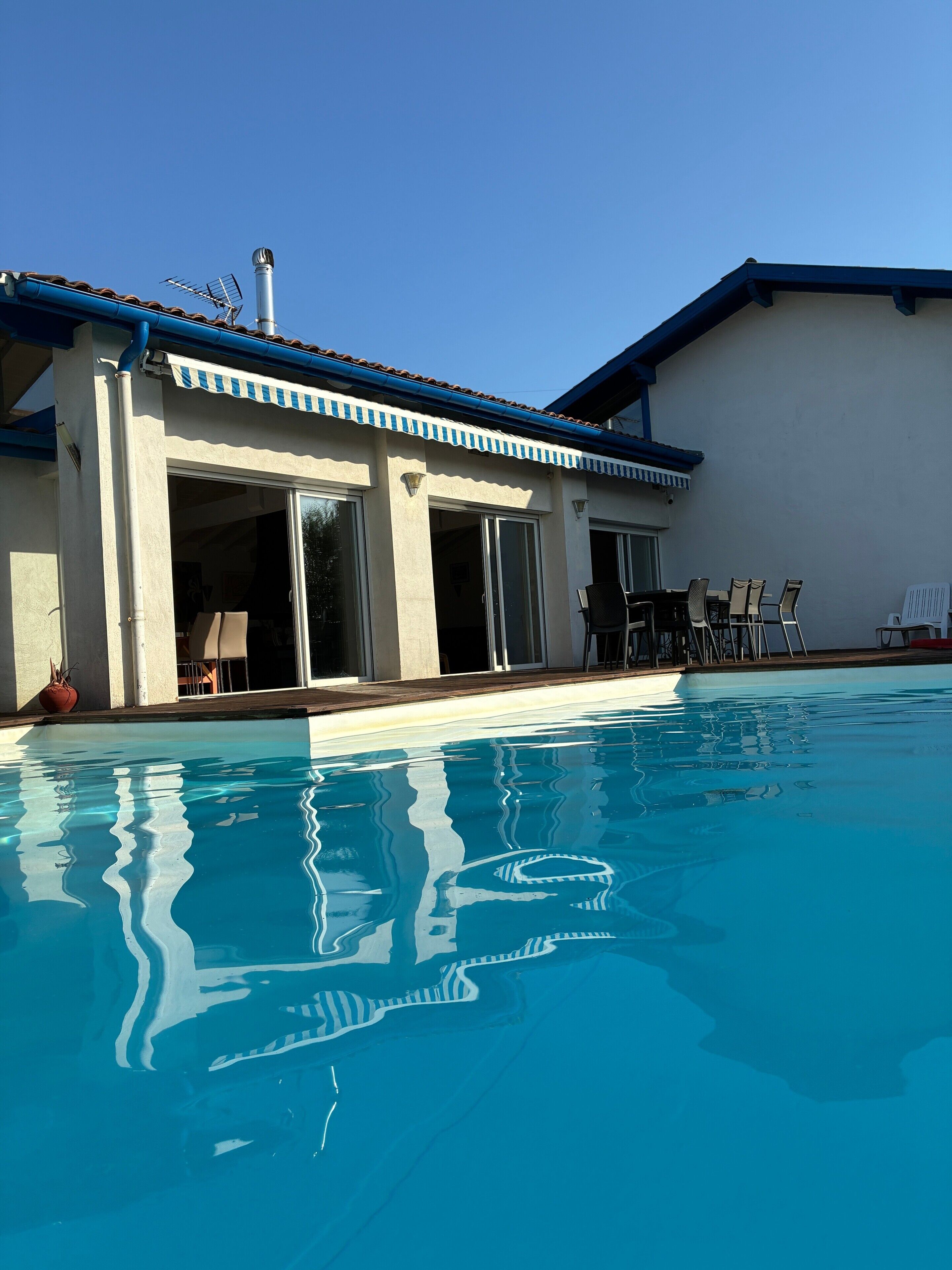 A swimming pool with a building adjacent to it, featuring a covered patio area with chairs and a table.