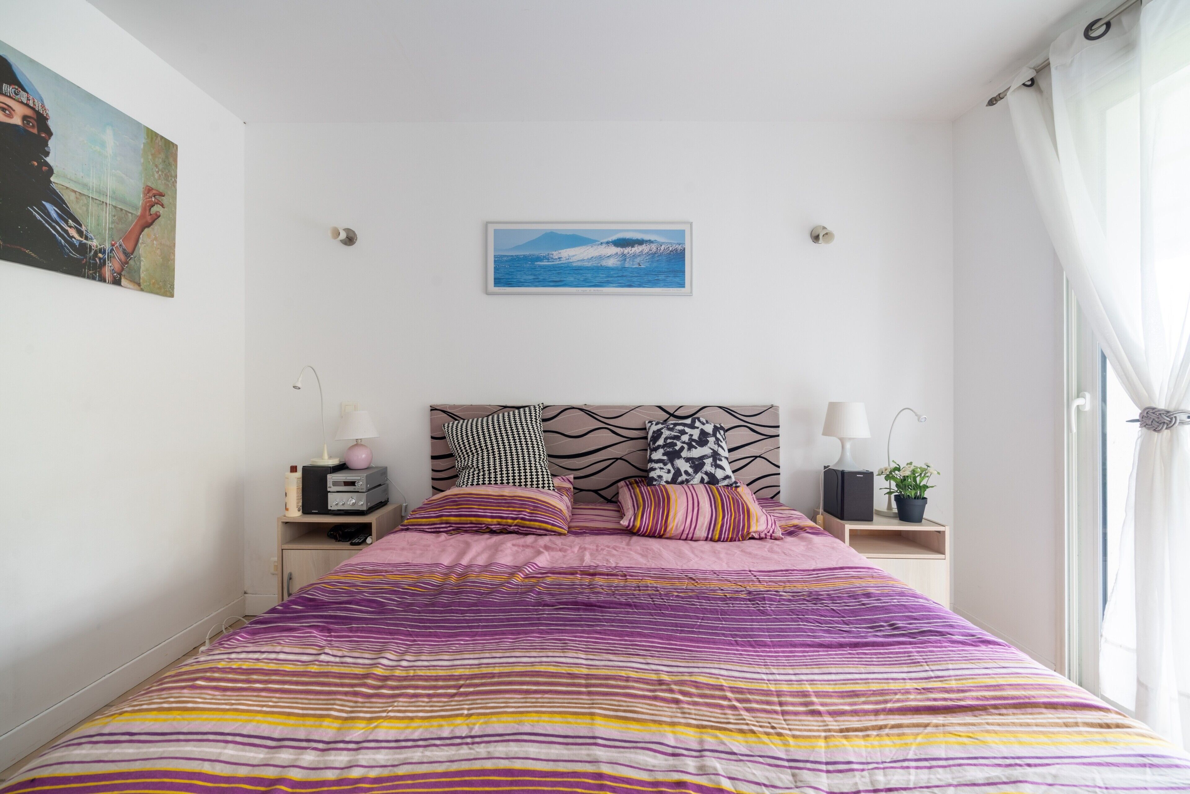 A bedroom with a bed, a headboard, a nightstand, a lamp, a plant, and a painting on the wall.