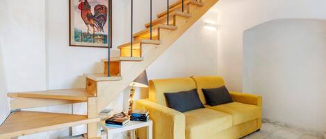 A staircase with wooden steps and black railings, a yellow sofa with dark cushions, a small white table with books, and a framed rooster painting on the wall.