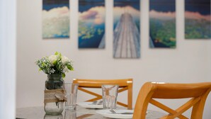 A dining table with a vase of flowers, clear glasses, and a plate with cutlery. There are wooden chairs and a wall with landscape paintings.