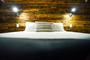 Una cama con sábanas blancas y almohadas a rayas, frente a una pared de madera con dos luces de pared.