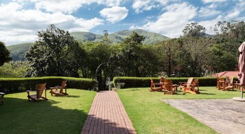 Un jardín con un camino pavimentado, sillas y mesas de madera, rodeado de exuberante follaje y colinas al fondo.
