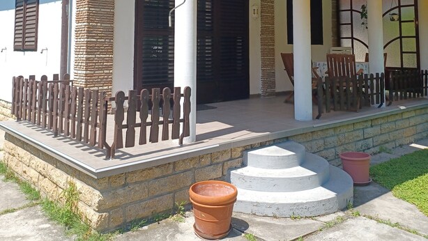 Una casa a due piani con un balcone, un portico con sedie e un piccolo giardino con una pianta in vaso.
