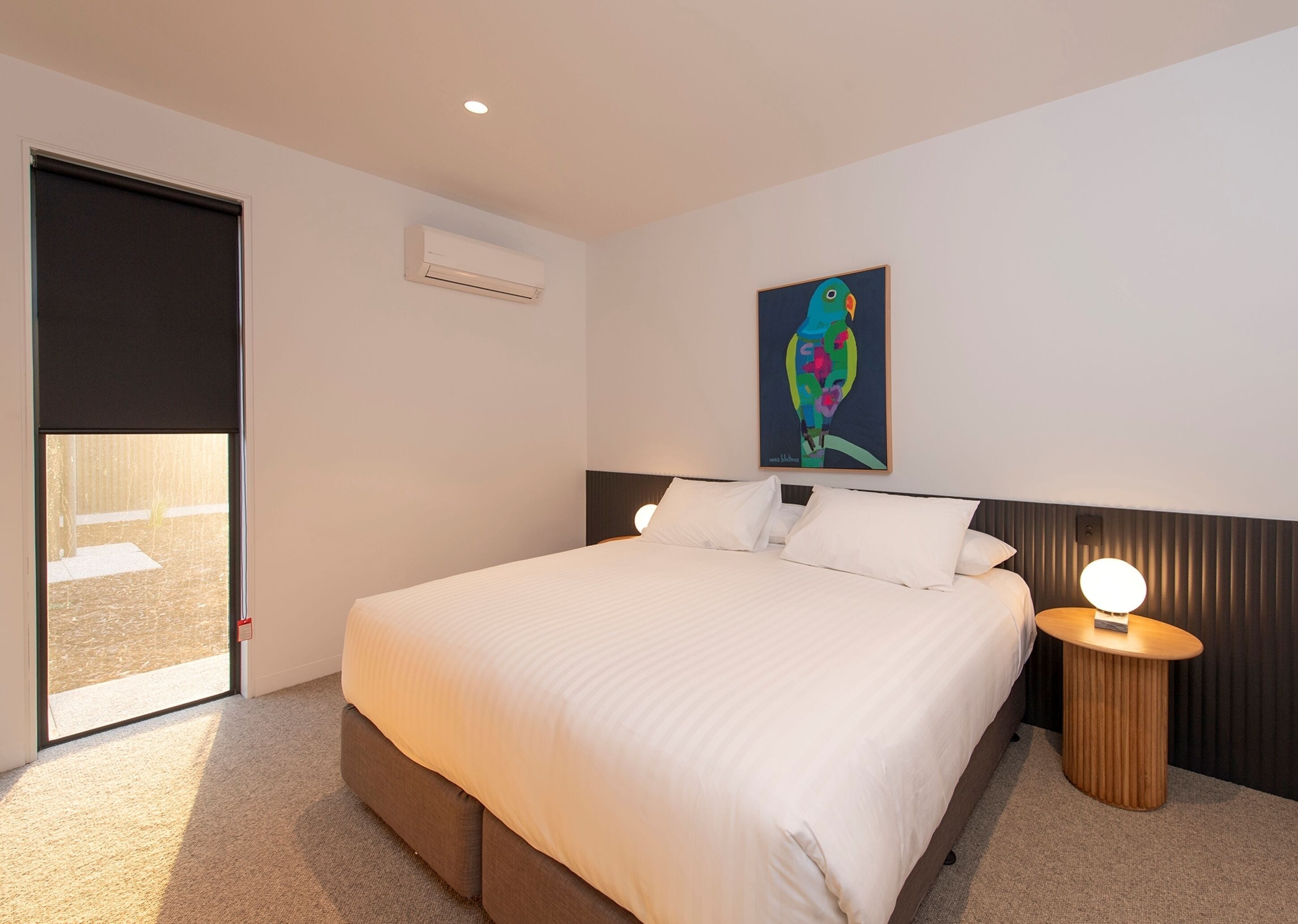 A bedroom with a bed, a nightstand, a wall-mounted air conditioner, a door, and a painting.