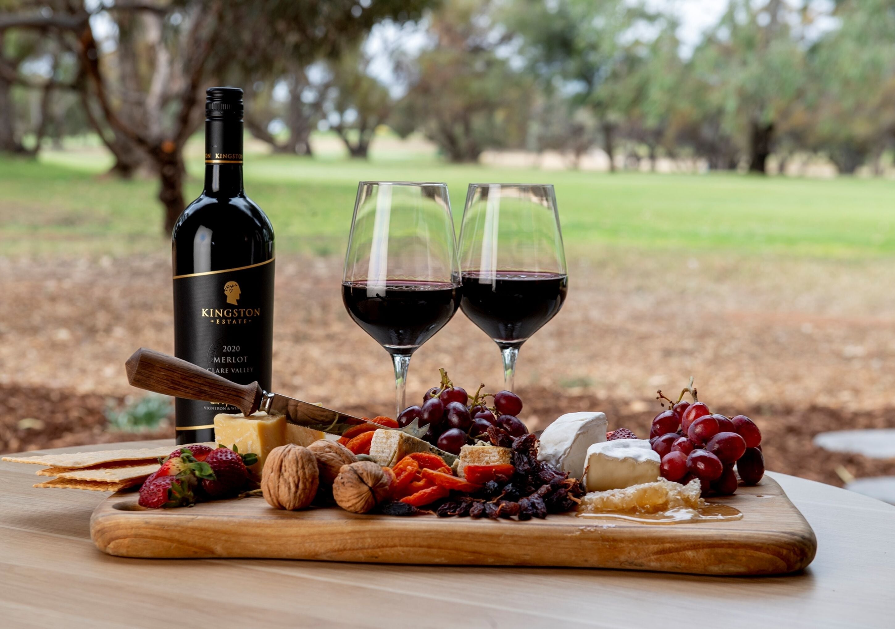 A wooden table with a bottle of wine, two glasses of red wine, and an assortment of cheeses, fruits, and nuts.
