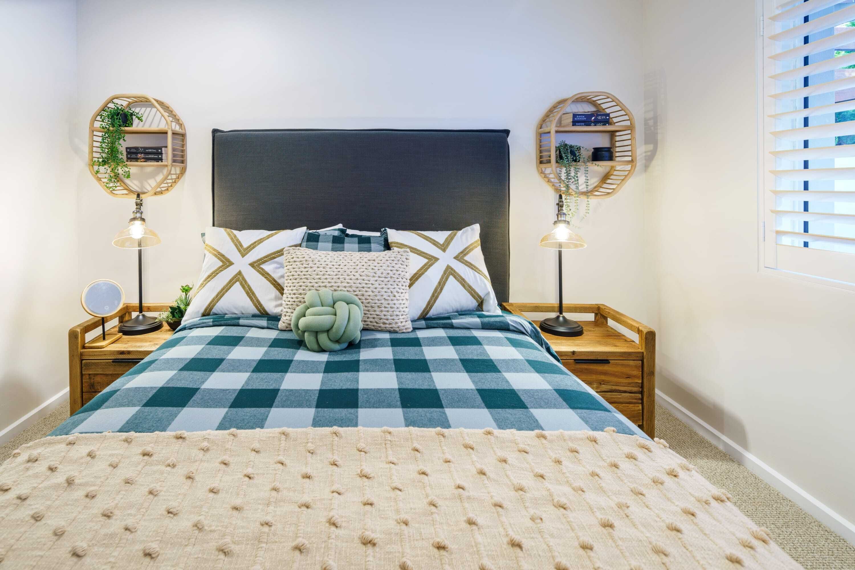 A bedroom with a checkered bedspread, a large headboard, two bedside tables with lamps, and a window with blinds.