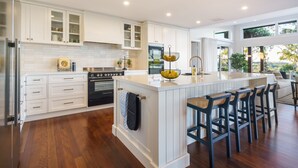 Una cocina moderna con armarios blancos, una isla central con taburetes y vistas al exterior a través de amplias ventanas.