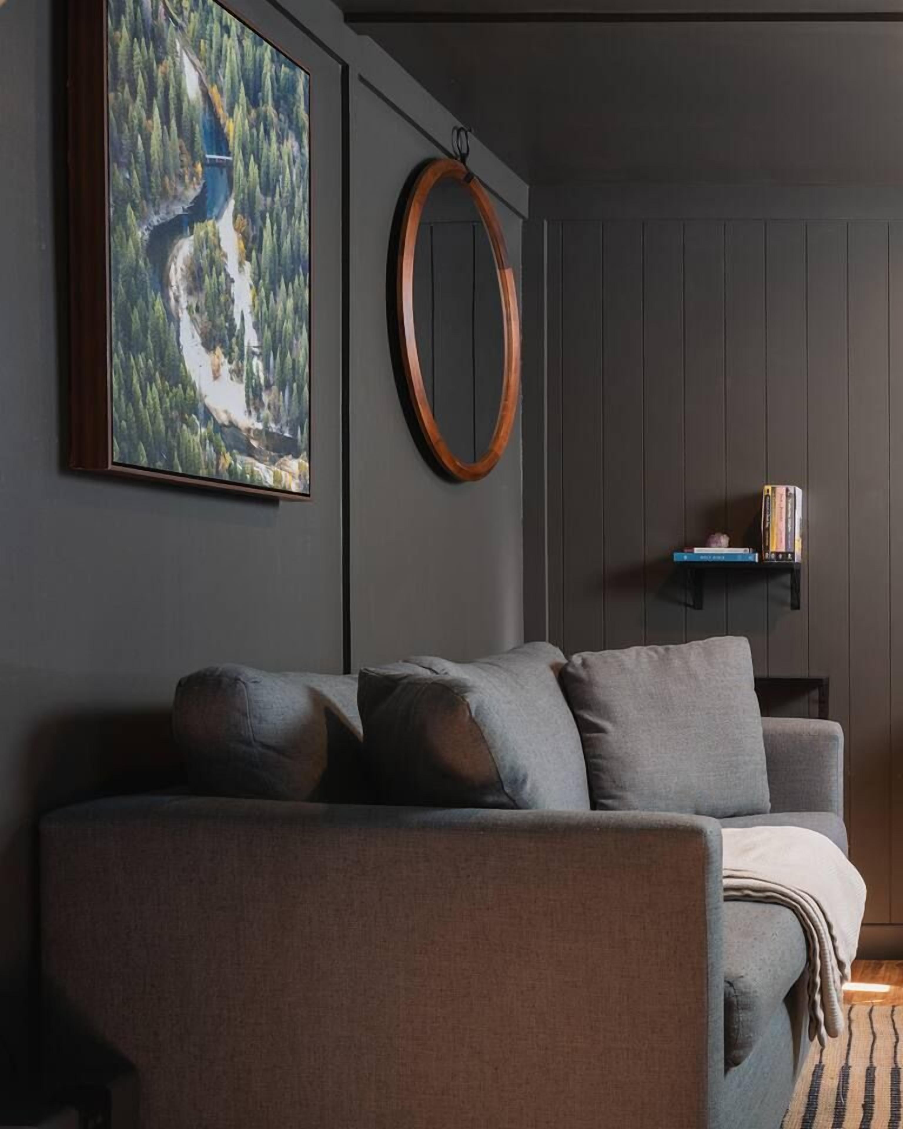 Una sala moderna con un sofá gris, un cuadro de un paisaje, un adorno circular de madera y una pequeña estantería.