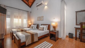 A hotel room with two beds, a wooden floor, a small rug, a desk, a chair, a mirror, and a trash bin. - Swarga Villa (Kandy)