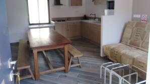 A kitchen with wooden cabinets, a sink, and a window. There is a wooden table and a bench. A white chair is visible in the foreground. - Gîte mon p'ti bout (LES ROCHES L'EVËQUE)