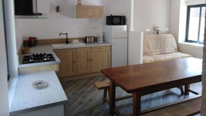 A compact kitchen with a stove, microwave, and sink. There is a wooden table and a couch in the adjacent living area. - Gîte mon p'ti bout (LES ROCHES L'EVËQUE)