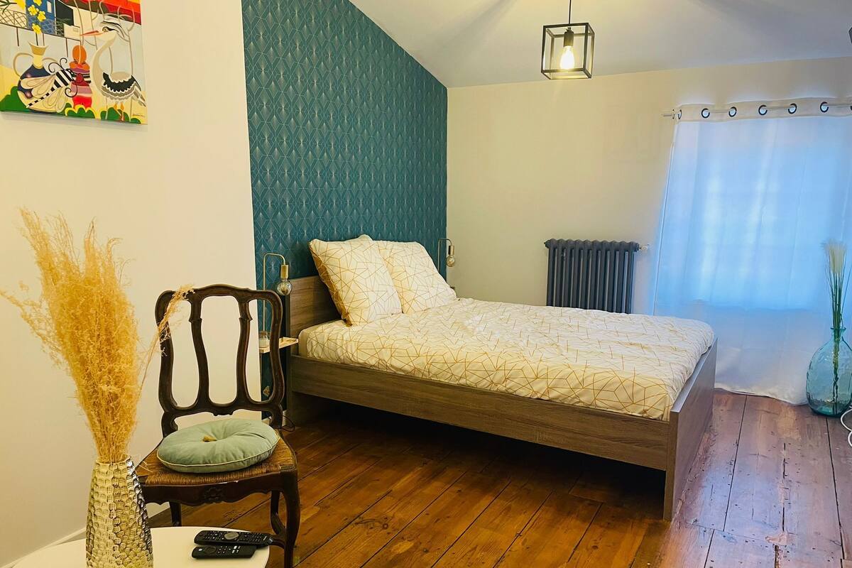 A bedroom with a wooden bed, a nightstand, a chair, a vase with dried plants, a remote control, and a painting on the wall.