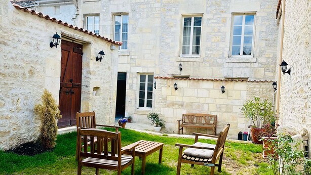 A stone building with a courtyard, featuring a wooden bench and chairs, and potted plants.