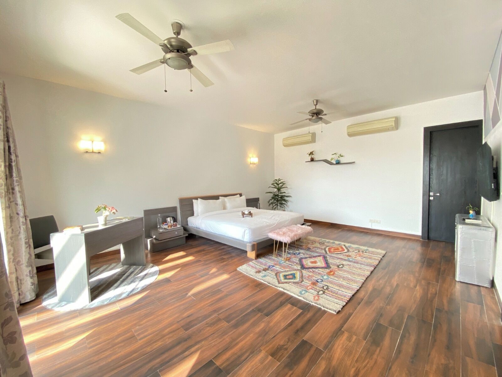Habitación de hotel con cama, escritorio, silla, dos ventiladores de techo, alfombra y una puerta.