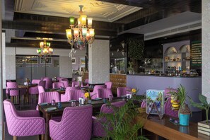 A restaurant with purple upholstered chairs, a chandelier, and a bar area with various items displayed. - Karma Lakelands (Gurugram)