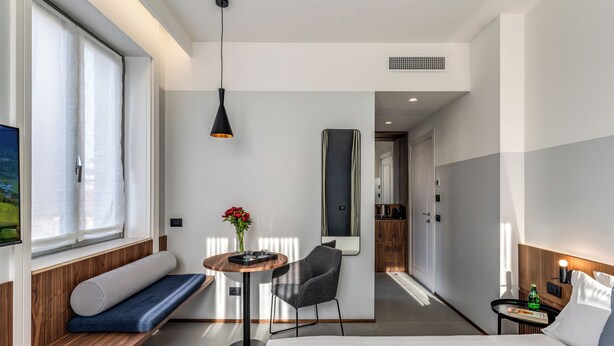 A modern hotel room with a bed, a seating area with a table and chair, a small table with a bottle, and a large window with curtains. - Sospeso Boutique Hotel (Milan)