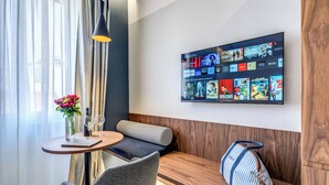 A hotel room with a wooden bench, a small table with a vase of flowers, a chair, and a wall-mounted TV displaying various streaming services. - Sospeso Boutique Hotel (Milan)