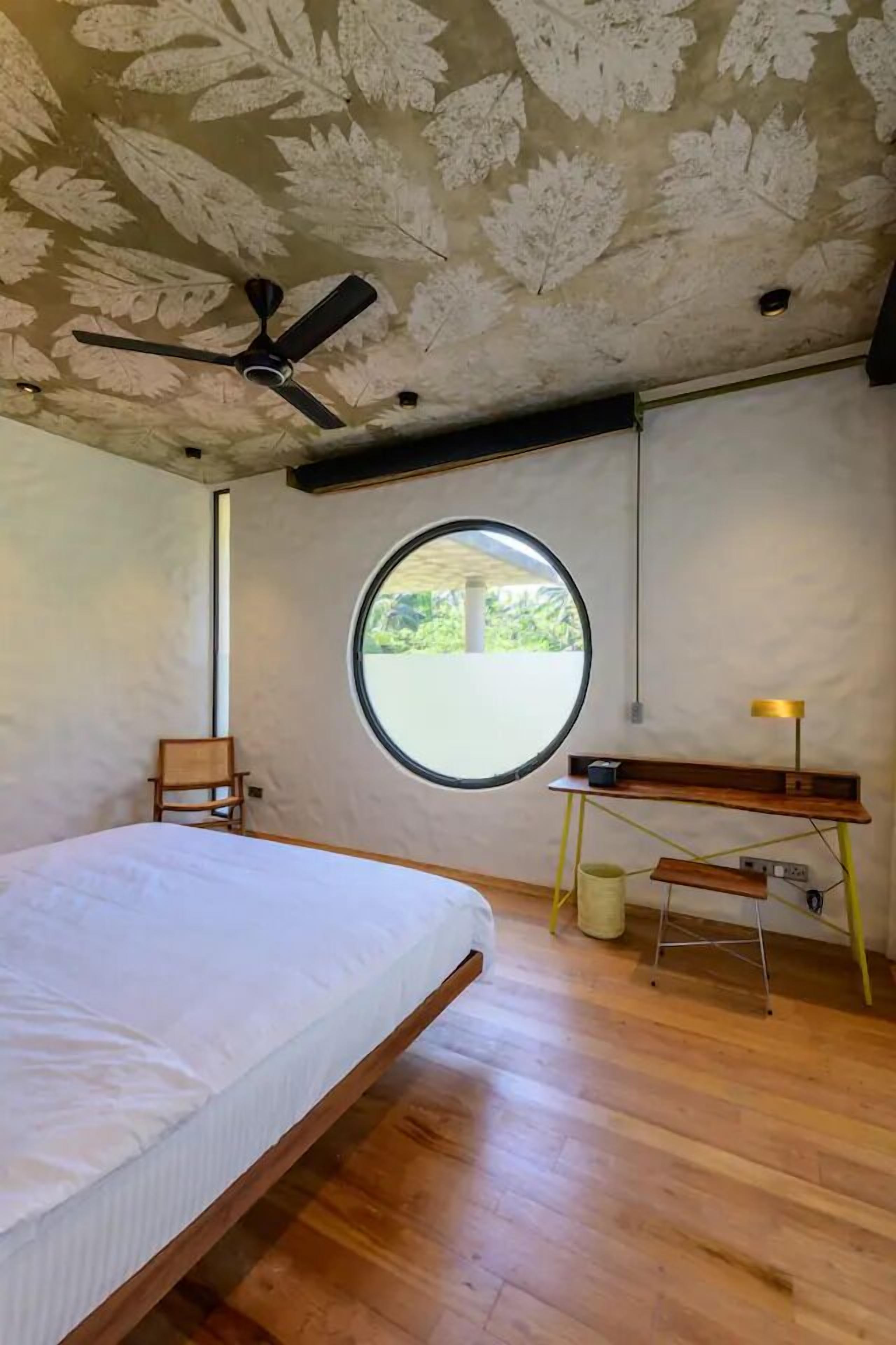 Un dormitorio con cama, ventilador de techo, ventana redonda, una silla de madera y un escritorio con lámpara.