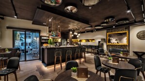Un área de bar moderna con barra central, diversas opciones de asientos incluyendo taburetes y mesas, y vista al exterior a través de un ventanal.