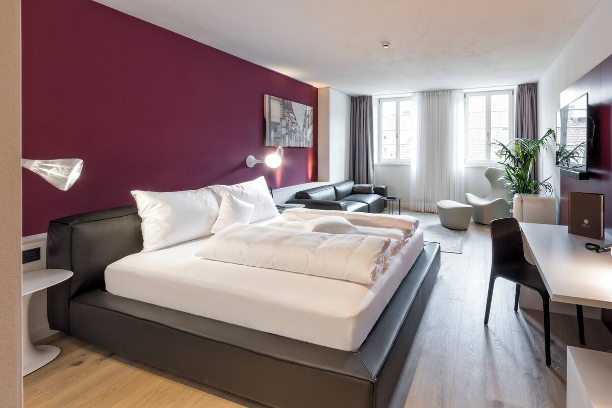 A modern hotel room with a large bed, a desk, a chair, and a sofa.