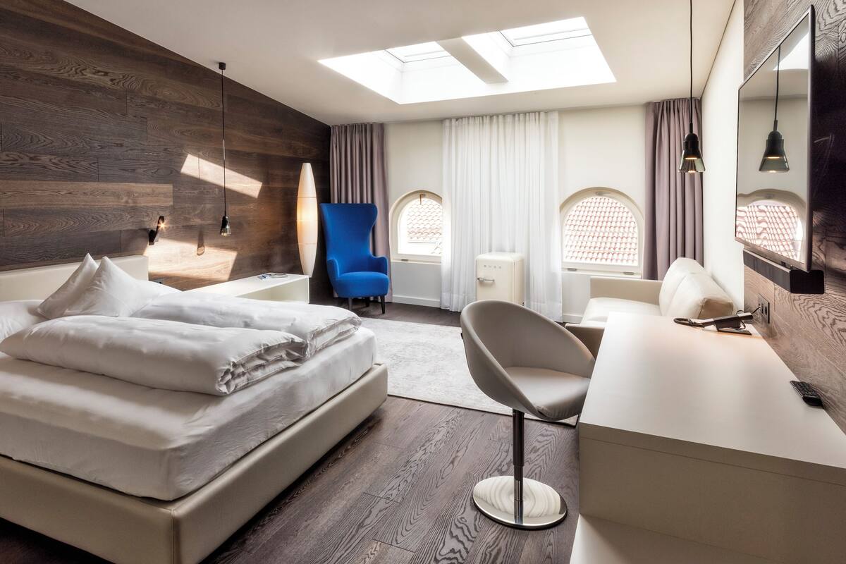 A modern hotel room with a large bed, a desk, a chair, and a small table. There are two windows with curtains, a skylight, and a wall-mounted TV.