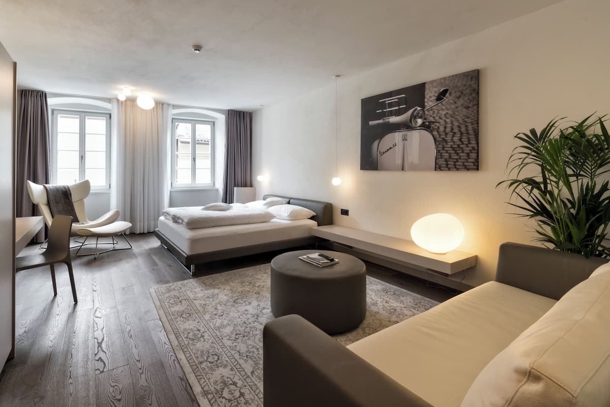 A modern hotel room with a bed, a seating area with a sofa and armchair, a small table, a large potted plant, and a wall-mounted artwork.