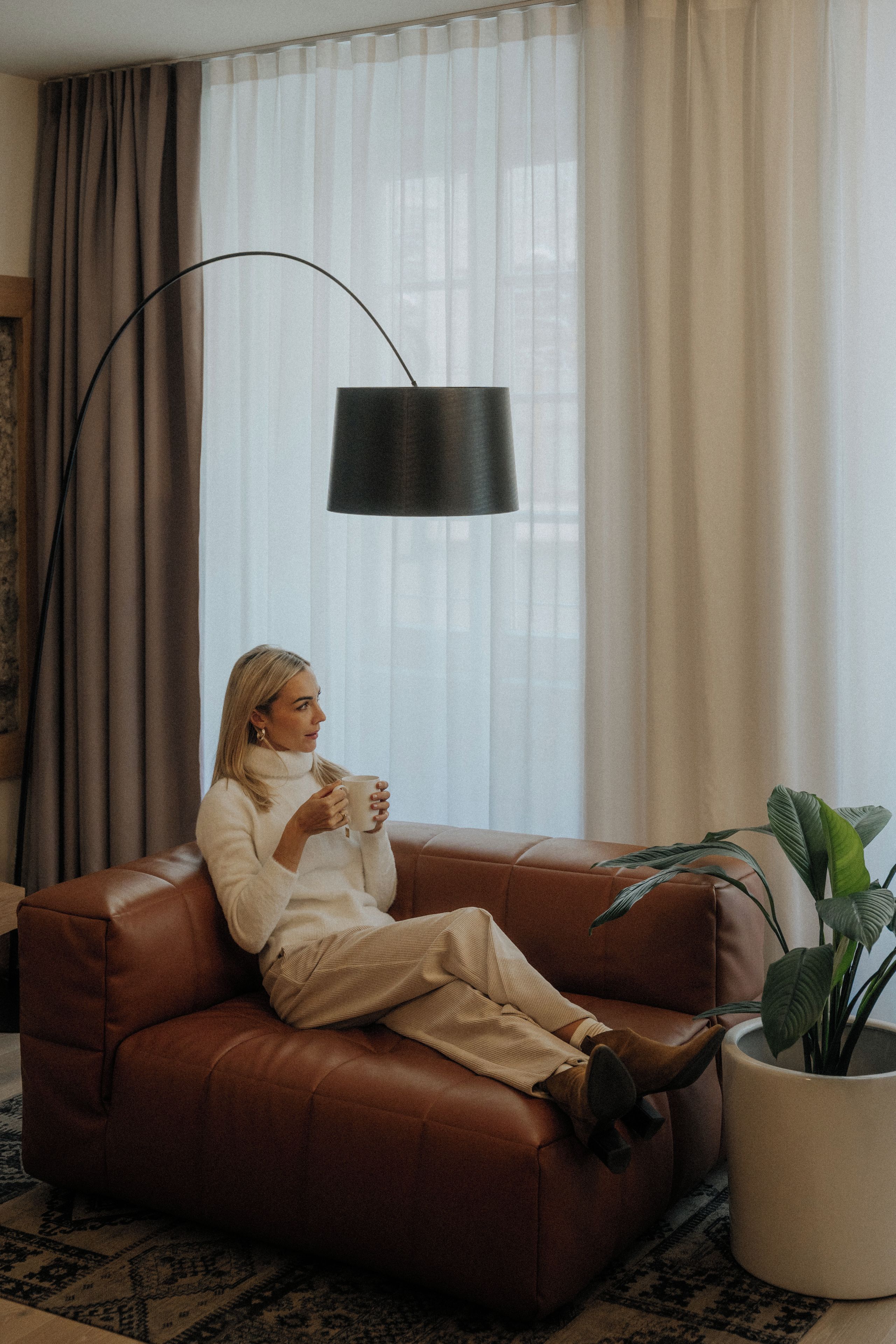 Una persona sentada en un sofá de cuero marrón, sosteniendo una taza, con una lámpara de pie y una planta en maceta en la habitación.