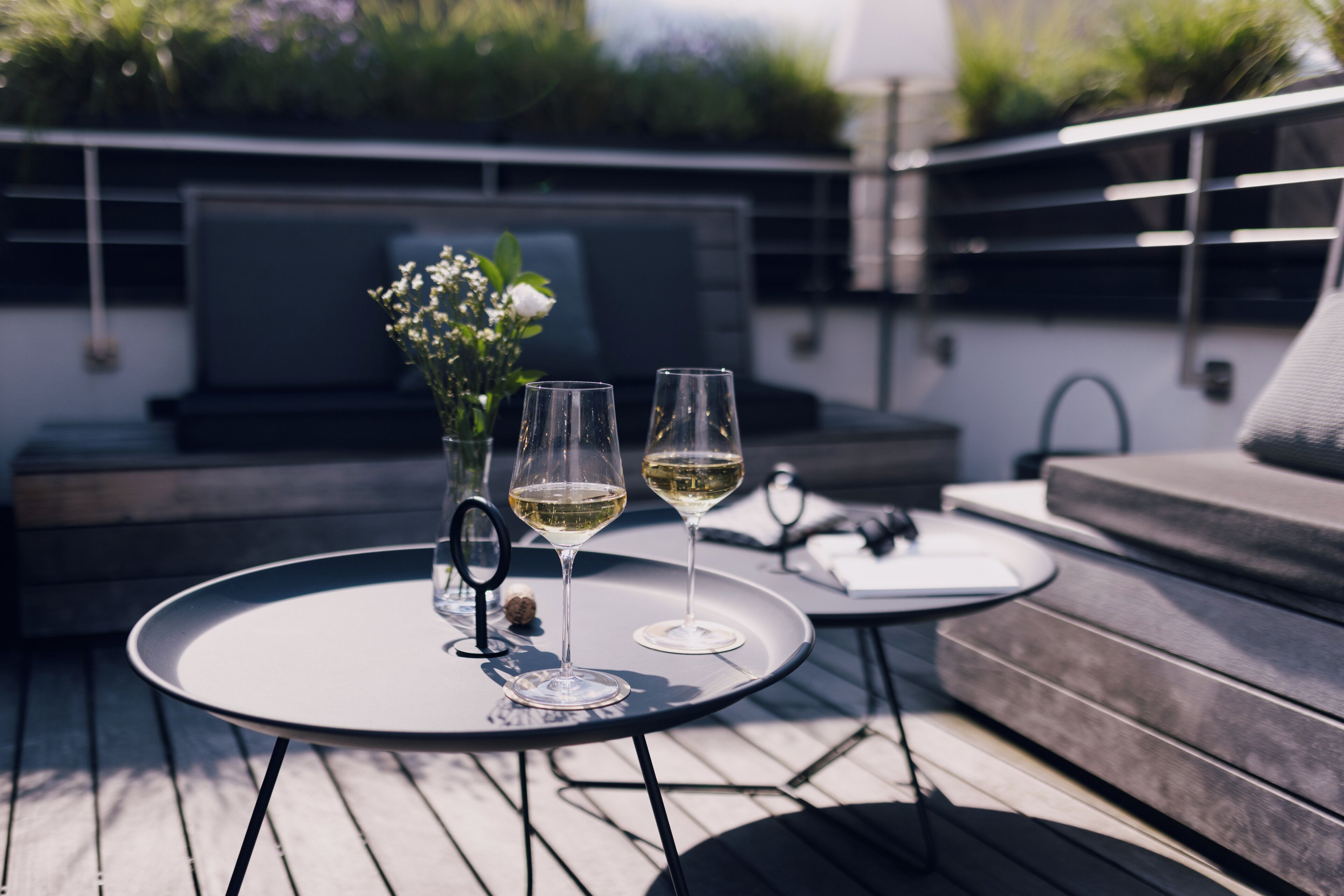 A round table with two wine glasses and a vase of flowers on a wooden deck.