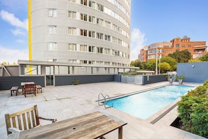 A rooftop swimming pool with a wooden bench and table, surrounded by a high-rise building with a modern design. - Auckland City Views Near Spark Arena Zodiak Stays (Auckland)