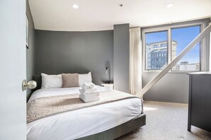 Una habitación de hotel moderna con una cama, una mesita de noche, una lámpara y vistas a la ciudad a través de un amplio ventanal.