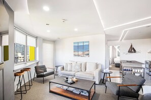 Una sala moderna con sofá, mesa de centro y sillones, una zona de cocina con mesa de comedor y un amplio ventanal con vista a la ciudad.