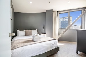 Moderni hotellihuone, jossa on suuri sänky, työpöytä ja ikkuna, josta avautuu näkymä kaupunkiin.