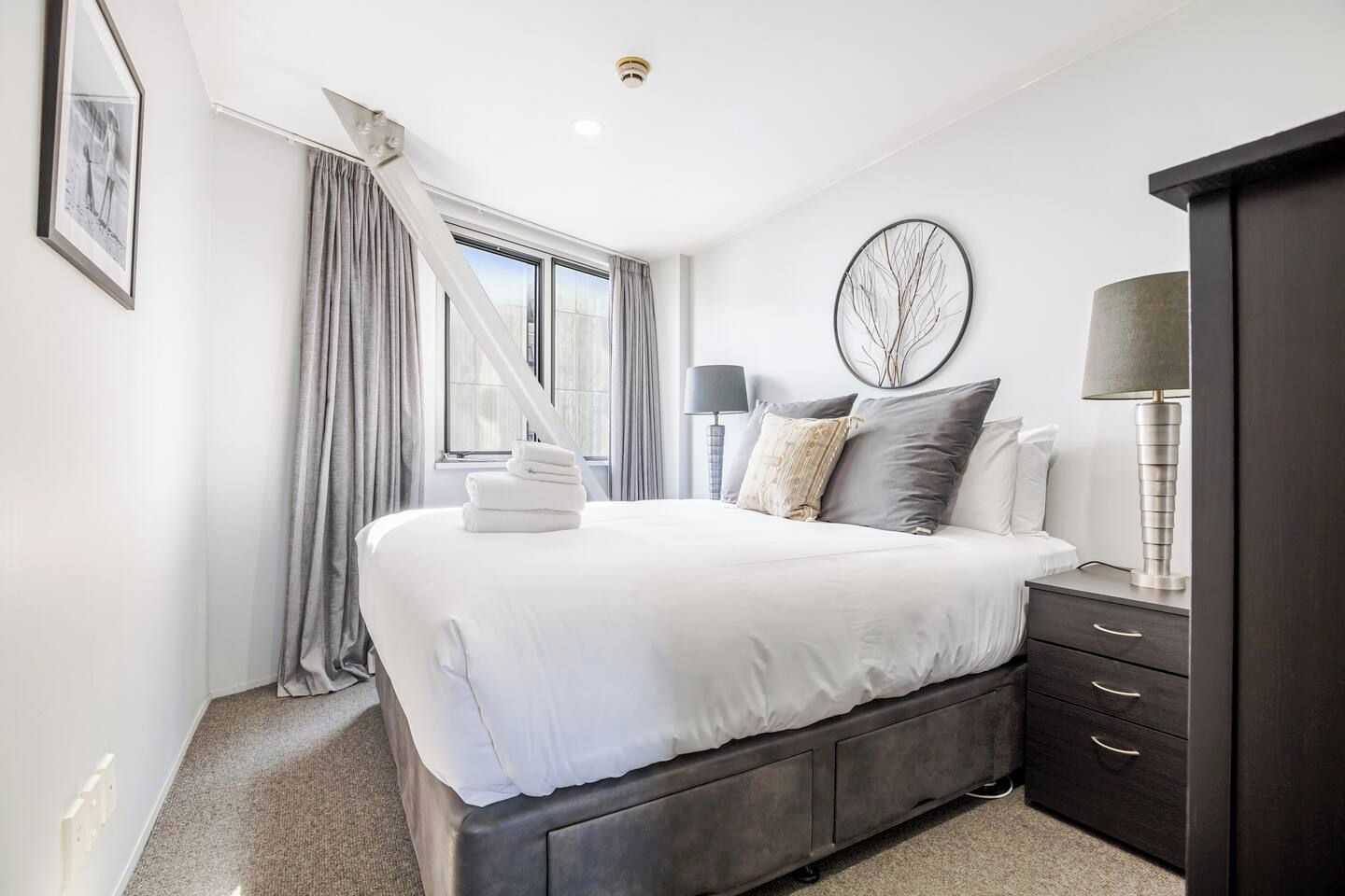 A modern bedroom with a large bed, a bedside table with a lamp, a dark wooden wardrobe, and a window with curtains.
