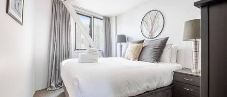 A modern bedroom with a large bed, a bedside table with a lamp, a dark wooden wardrobe, and a window with curtains.