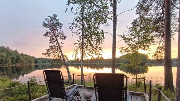Een houten terras met twee stoelen en een tafel, met uitzicht op een rustig meer bij zonsondergang.